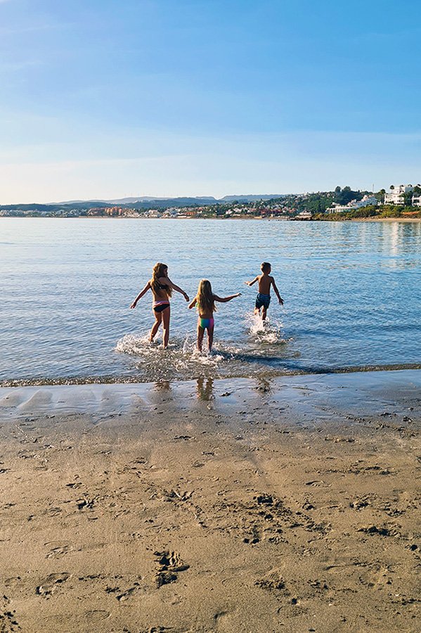 Drie kinderen rennen met enthousiasme het water in op het strand van Playa del Cristo, omgeven door gouden zand en turquoise water onder de warme middagzon.