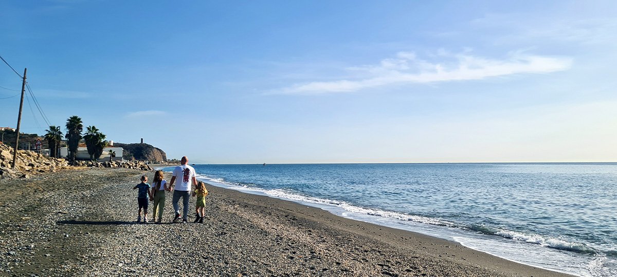 Drie kinderen lopen hand in hand met hun vader op het strand van La Cala Del Moral, omgeven door de warme gloed van de ondergaande zon.