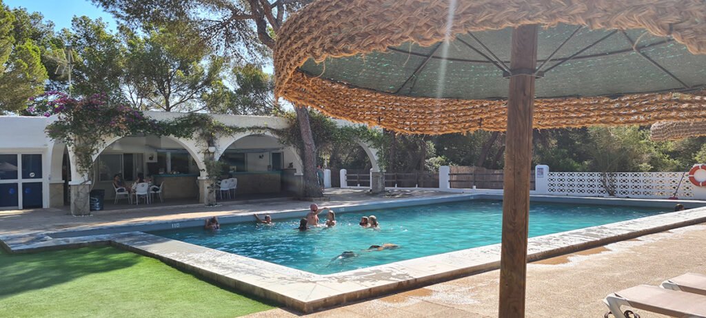 Een levendig zomer tafereel bij een resort zwembad, omlijst door een parasol van gevlochten stro op de voorgrond, met mensen die zwemmen en ontspannen op ligstoelen.