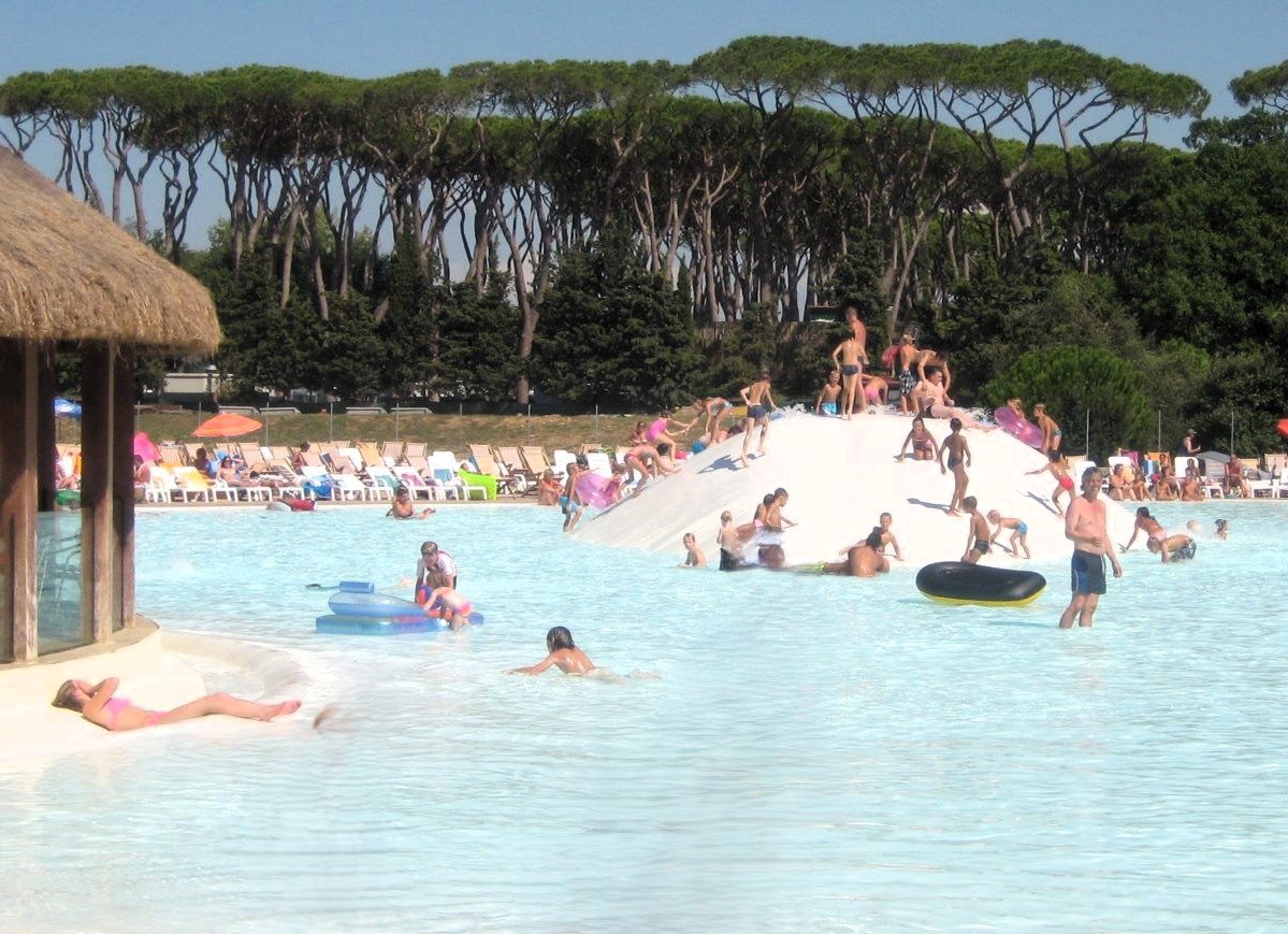 Kinderen en volwassenen genieten van een grote glijbaan en verkoelend water in het zwembad van Camping HU Park Albatros Village, omgeven door de unieke mediterrane pijnbomen van San Vincenzo.